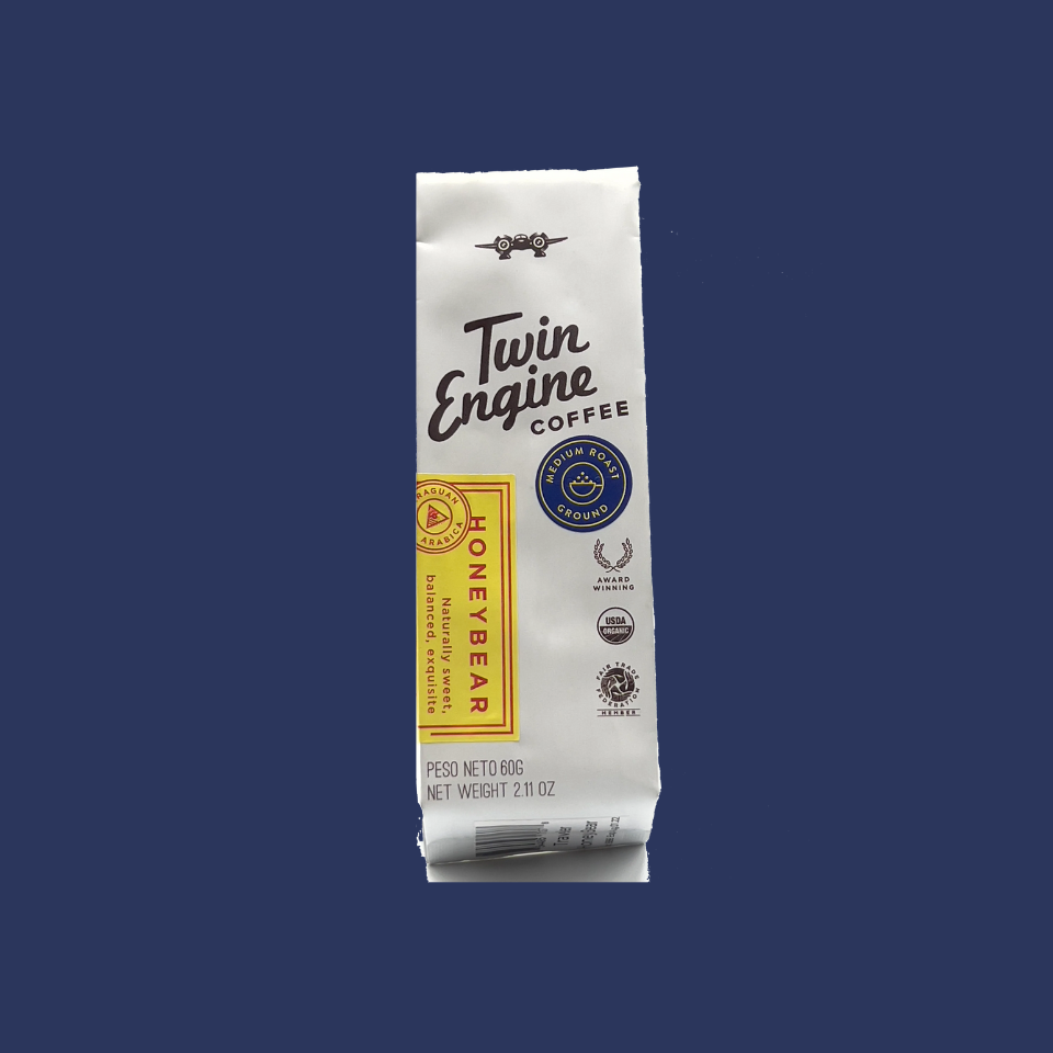 Coffee grounds package labeled Twin Engine Coffee Honey Bear flavor; placed on a blue background
